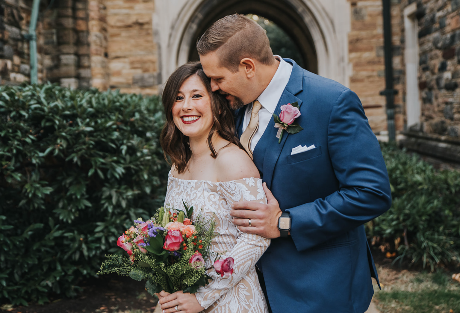 Elopement Wedding A bride in an off-shoulder white lace dress holds a vibrant bouquet as she smiles joyfully. A groom in a blue suit, white shirt, and rose boutonniere stands beside her, whispering into her ear. They are posed in front of a stone archway, with lush greenery surrounding them. Elopements Inc